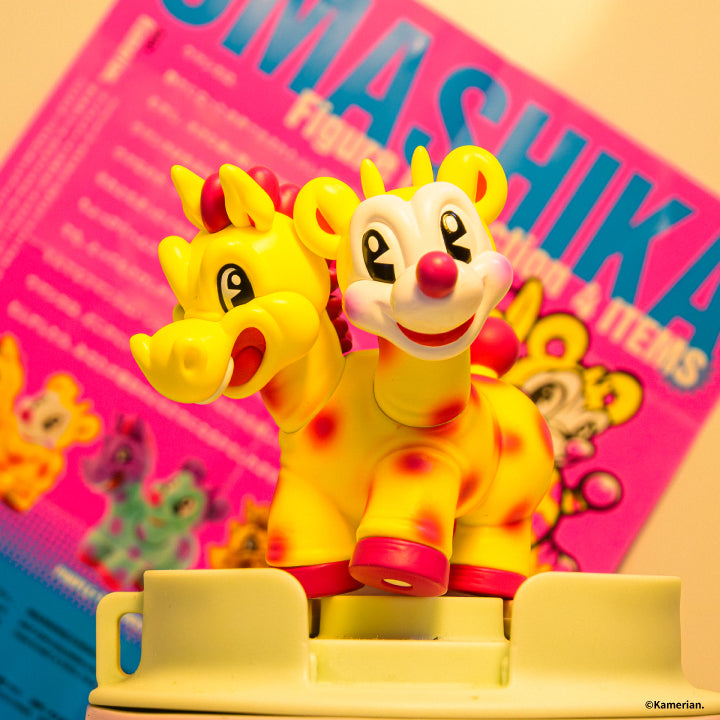 Yellow UMASHIKA figure displayed in front of colorful UMASHIKA artwork