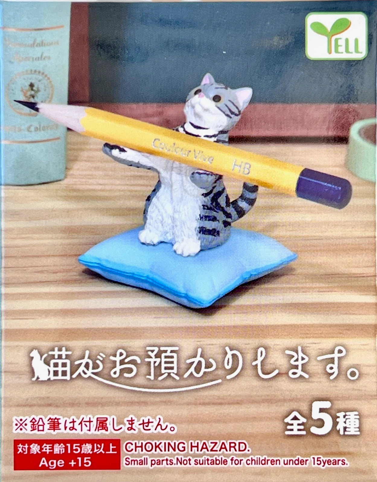 A small figurine of a gray tabby cat sitting on a blue cushion and holding a yellow pencil, shown on a wooden tabletop with Japanese text and a YELL brand mark in the top right.